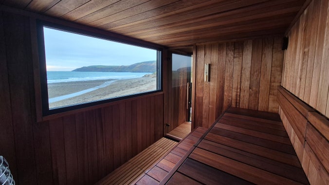 Golygfa o'r traeth a'r môr drwy ffenestr sawna bren, wedi'i leoli ym Mhorth y Swnt.